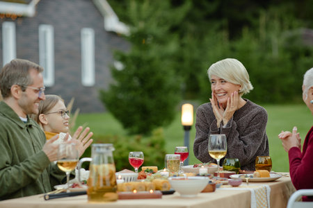 Caucasian senior woman smiling with hands on face while sitting at outdoor table with multiethnic family including middle aged man, senior man, and young girl enjoying meal togetherの写真素材