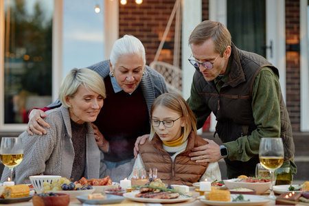 Caucasian senior woman, middle aged man, middle aged woman and preteen girl gathering around outdoor table celebrating birthday with cake and candles, sharing family momentの写真素材