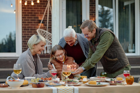 Middle aged man presenting birthday cake with candles to smiling girl, surrounded by middle aged woman and senior woman during outdoor family dinner celebrationの写真素材