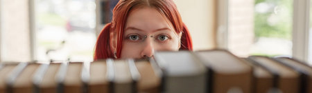 Portrait of Caucasian teenage girl with red hair looking directly at camera through row of books on shelf, eyes visible above books, standing in library or bookstore settingの写真素材