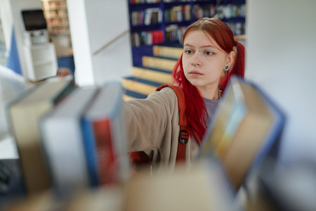 Caucasian teenage girl with red hair reaching for book on library shelf, focused expression on face, surrounded by blurred books in foregroundの写真素材
