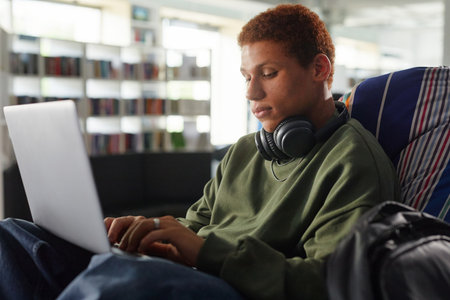 Young man sitting in modern library using laptop, focusing on typing, studying or working on digital project in relaxed environmentの写真素材