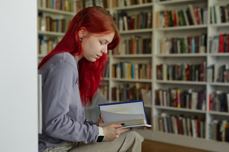 Teenage girl with long red hair sitting in library reading book, holding open hardcover while focusing on pages, surrounded by shelves filled with books in backgroundの写真素材