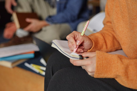 Girl writing in notebook with pen, sitting indoors with other people in background, focusing on taking notes during group activity or meetingの写真素材