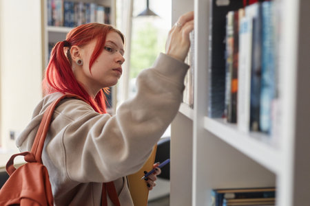 Teenage Caucasian girl with red hair browsing bookshelf, holding notebook and pen, wearing backpack, standing in library or bookstore, searching for book, focused expressionの写真素材