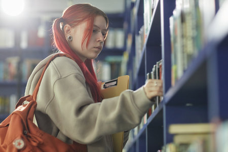 Teenage Caucasian girl with red hair searching books on library shelf, holding folder and backpack, focused expression, standing in academic environment, side profile visibleの写真素材