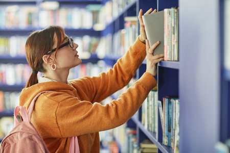 Caucasian young adult woman with glasses reaching for book on library shelf, wearing backpack, standing in front of rows of books, engaging in academic or leisure reading activityの写真素材