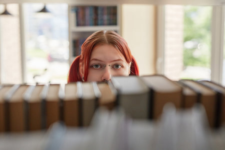 Portrait of girl with red hair looking through bookshelf, eyes visible above row of books, standing in library or bookstore, natural light coming from windowの写真素材