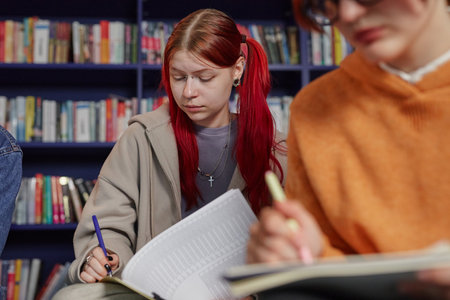 Teenage girl with red hair sitting in library studying and writing in notebook, looking down at paper, another young adult woman with glasses in foreground also writingの写真素材