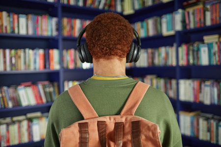 Back view of Black teenager wearing backpack and headphones standing in front of bookshelves in library, appearing to listen to audio or music while looking at book selectionの写真素材