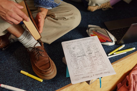 Young adult man sitting on floor holding book near handwritten notes, colored markers and open laptop, studying or preparing for exam in casual indoor settingの写真素材