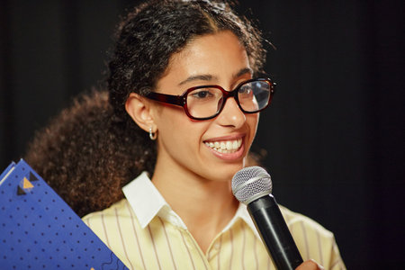 Portrait of young adult biracial woman wearing glasses smiling and speaking into microphone while holding folder, curly hair pulled back, standing against dark backgroundの写真素材