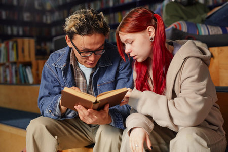 Teenage girl with red hair and boy sitting together reading book, both focused on pages, sharing learning experience in library settingの写真素材