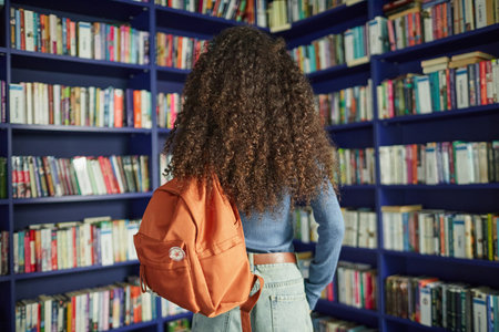 Teenage girl standing with backpack in front of bookshelves in library, facing away from camera, long curly hair visible, exploring book selection, casual clothingの写真素材