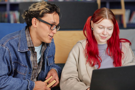 Young adult Asian man and young adult Caucasian woman sitting together using laptop, man holding...
