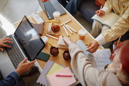 Group of young adult multiethnic students collaborating around table, using laptop, writing in notebooks, sharing muffins, discussing ideas, working together on creative projectの写真素材