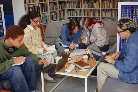 Group of multiethnic teenagers collaborating on project, using laptops and writing in notebooks while sitting around table in library, focused on studying and sharing ideas togetherの写真素材