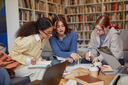 Three young adult girls sitting together studying and writing in notebooks, collaborating on academic project in library with laptops and coffee cups visibleの写真素材