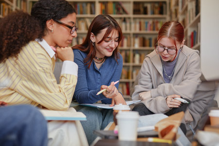 Group of multiethnic teenage girls collaborating on school project in library, discussing notes and studying together with books and stationery on table, focused on teamwork and learningの写真素材