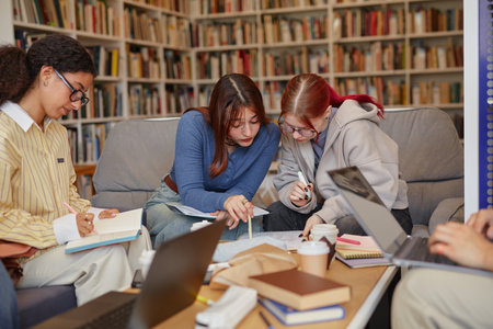 Group of multiethnic teenagers collaborating on school project, sitting together on couch in library, writing notes and using laptops, focused on teamwork and studying for academic assignmentの写真素材