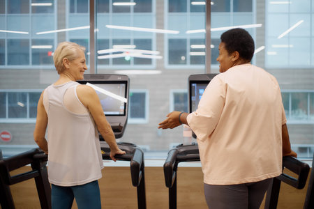Caucasian senior woman and Black middle aged woman using treadmills in modern gym, both standing...