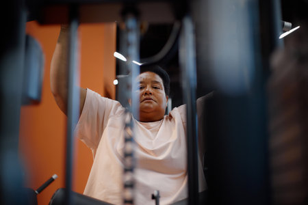 Middle aged Black woman exercising on gym machine, lifting arms while focusing on workout, visible determination on face, fitness equipment framing subject in modern gym settingの写真素材