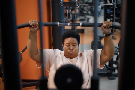 Portrait of middle aged Black woman exercising on strength training machine in gym, gripping handles and lifting weights, focused facial expression, fitness equipment visible in backgroundの写真素材