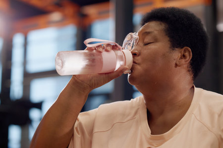 Middle aged Black woman drinking from water bottle, sitting indoors in gym or fitness center,...