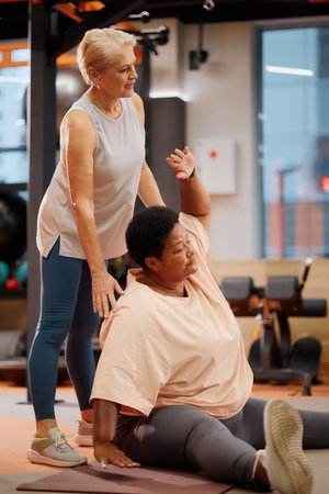Aged woman assisting woman stretching on exercise mat in gym, both focusing on flexibility training,...