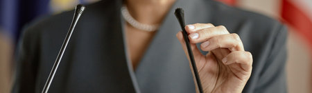Header of woman standing at podium adjusting microphone with right hand, preparing for public speech or press conference, wearing formal business attire, flags in backgroundの写真素材