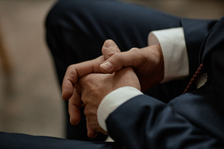 Man clasping hands together while sitting in formal attire, fingers interlaced and resting on lap, close up of hands and part of suit visibleの写真素材