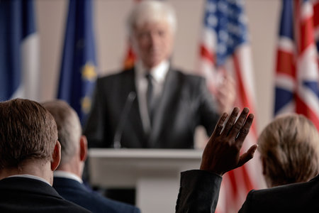 Caucasian senior man speaking at podium during press conference, audience members raising hands to ask questions, multiple national flags visible in backgroundの写真素材
