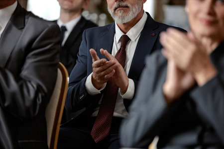 Middle aged Caucasian man in suit sitting among group of professionals clapping hands during formal event, surrounded by other adults in business attire, focus on hands and gestureの写真素材