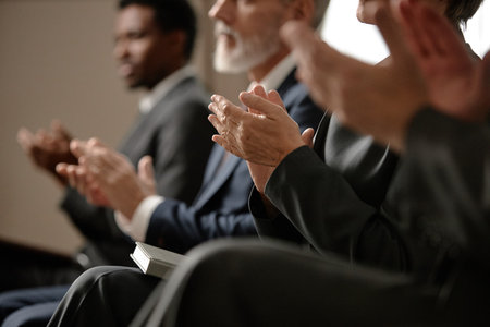 Group of middle aged and senior men and women of diverse ethnicities sitting in row clapping hands during event, focus on hands and partial profiles, business attire visibleの写真素材