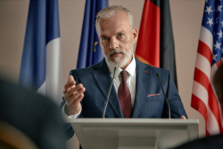 Portrait of senior Caucasian man speaking at podium gesturing with hand during press conference with international flags in background, maintaining serious facial expressionの写真素材