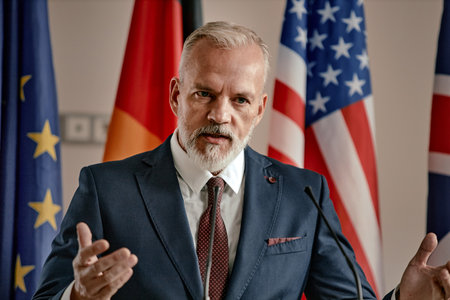 Portrait of middle aged Caucasian man speaking at podium gesturing with hands, standing in front of European Union, German, and United States flags during press conferenceの写真素材
