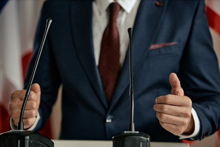 Man gesturing with hands while standing behind podium with microphones, wearing formal suit and tie, delivering speech or addressing audience at eventの写真素材