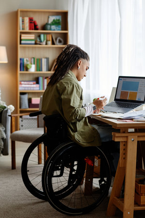 Young adult woman with disability sitting in wheelchair working at desk using laptop and writing notes in home office setting, bookshelf and window in backgroundの写真素材