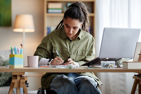Young adult woman with disability sitting in wheelchair writing notes at desk with laptop and stationery, focusing on studying or working in modern home office environmentの写真素材