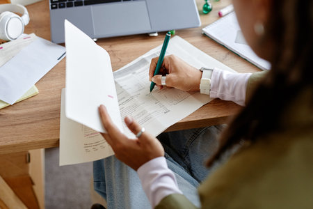 Young adult woman reviewing financial documents while writing notes at desk, holding paperwork in...