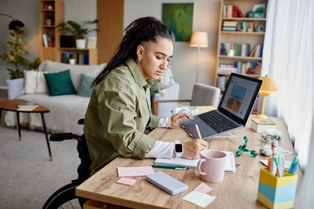Young adult woman with disability sitting in wheelchair working at desk using laptop and writing in...