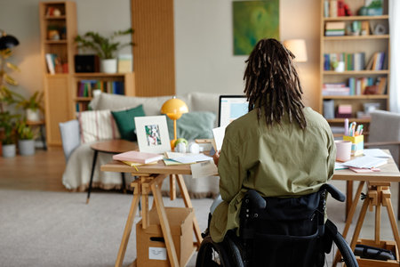 Young adult woman with disability sitting in wheelchair working at desk with computer in modern home office, surrounded by documents and personal items, facing away from cameraの写真素材