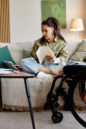 Young adult woman with disability sitting cross legged on sofa reviewing documents and looking at digital tablet, wheelchair visible in foreground, casual home settingの写真素材