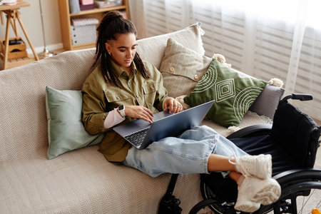 Young adult woman with disability sitting on sofa using laptop, resting legs on cushions, working or studying remotely, wheelchair visible beside, focused on screenの写真素材