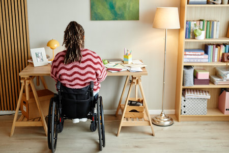 Woman with disability sitting in wheelchair working at wooden desk in home office, surrounded by books and stationery, long braided hair visible from behind, casual indoor settingの写真素材