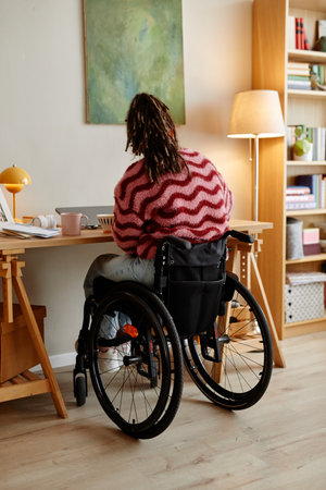 Young adult woman with disability sitting in wheelchair working at desk in home office, facing...