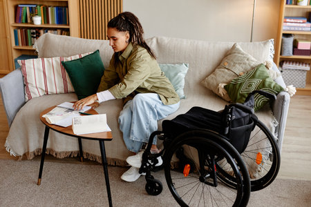 Young adult woman with disability sitting on sofa sorting through paperwork on small table, wheelchair positioned nearby, focused on organizing documents in living room settingの写真素材
