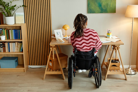 Young adult woman with disability sitting in wheelchair working at desk in home office, back view showing workspace with documents and stationery, focused on task completionの写真素材