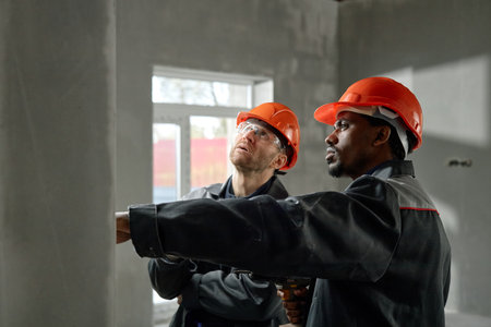 Caucasian middle aged man and Black middle aged man wearing safety helmets inspecting construction site, standing indoors with one pointing and discussing project detailsの写真素材