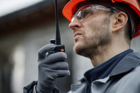 Caucasian middle aged man wearing safety helmet and protective glasses holding walkie talkie, communicating during construction work, close up of face and hand, looking awayの写真素材